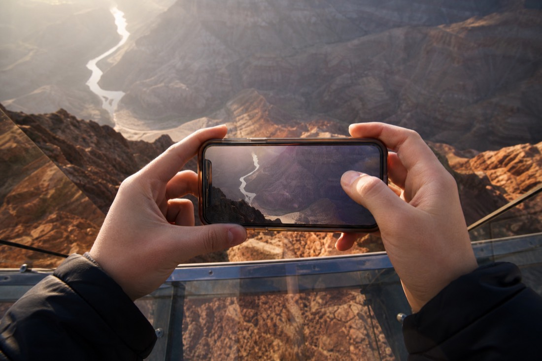 A visitor's hands holding a phone up over a glass walkway with the canyon floor visible far below