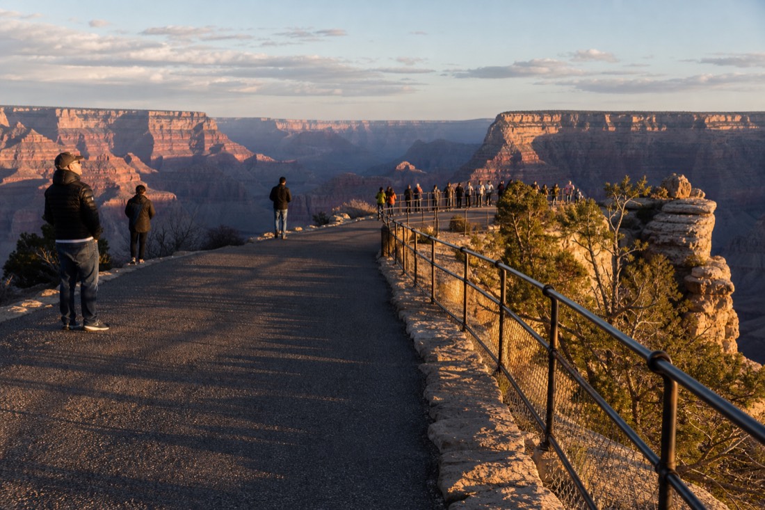Visitors spread along the paved South Rim promenade at golden hour with the canyon glowing behind the railing