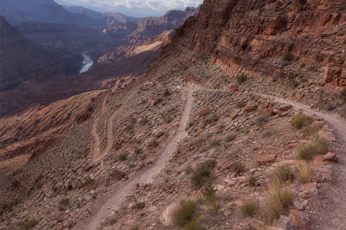 A steep inner-canyon trail switchbacking down red cliff bands toward the distant Colorado River