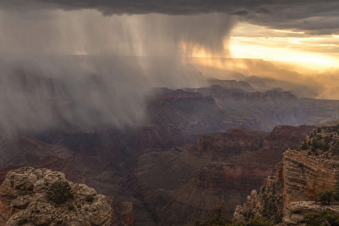 A dark monsoon thunderstorm with curtains of rain moving across the Grand Canyon while sunlight breaks through behind it