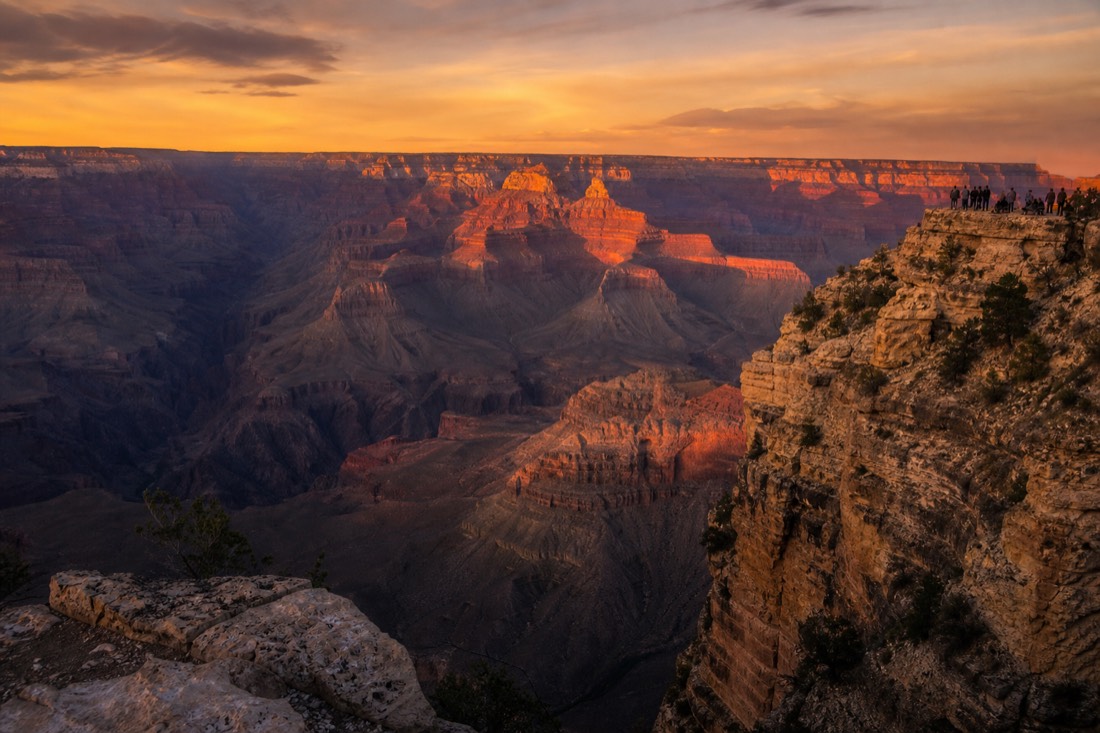 Sunset light turning the Grand Canyon walls deep orange and purple as small groups watch from the rim