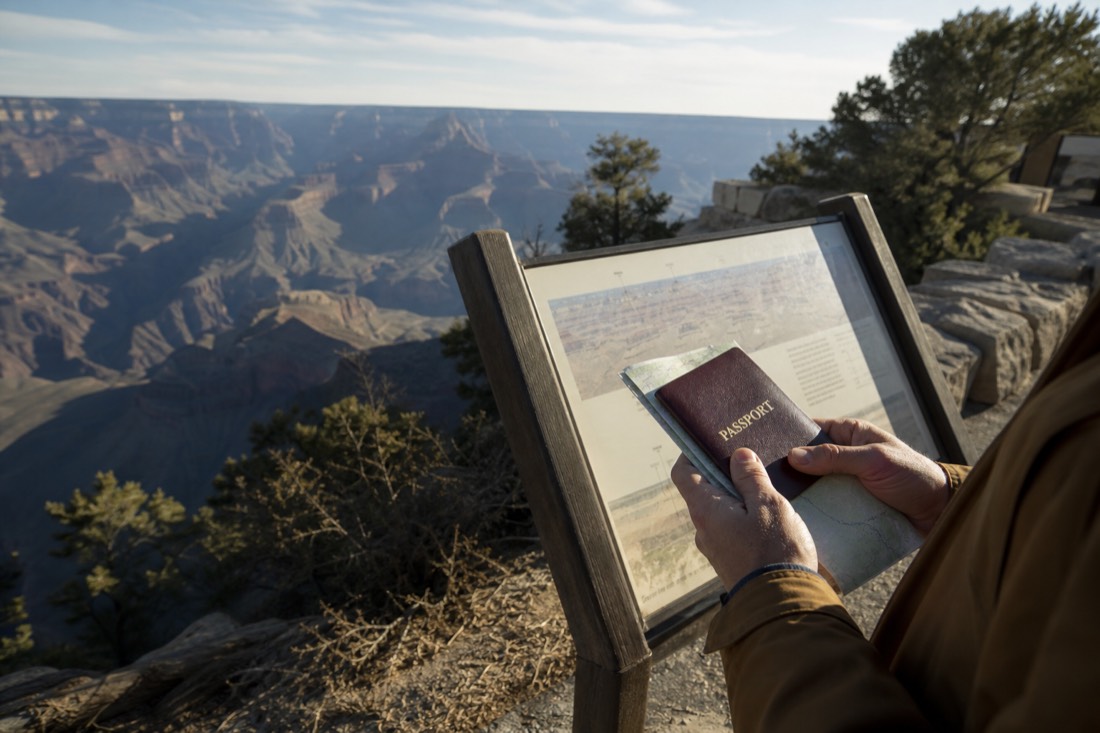A traveler holding a passport and park map at a wooden information board near the Grand Canyon visitor center