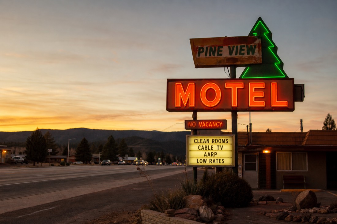A warm-lit roadside motel sign at dusk in Williams Arizona with pine ridgelines behind the highway