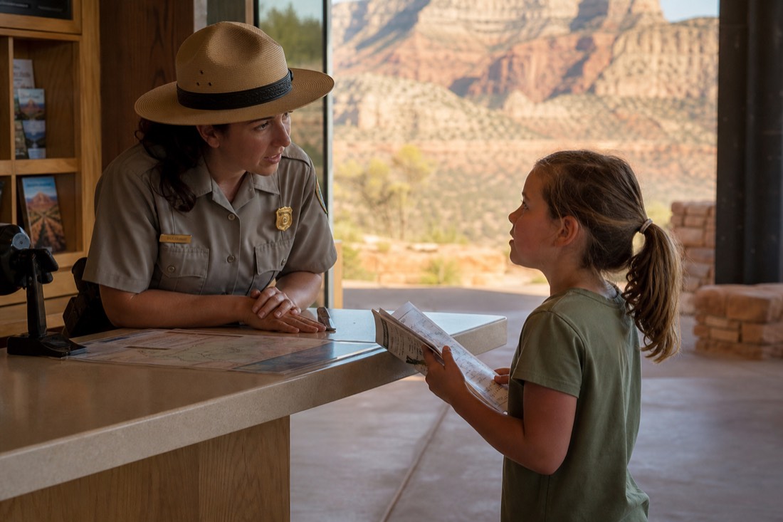 A park ranger in a flat hat talking with a child holding an activity booklet near a visitor center desk