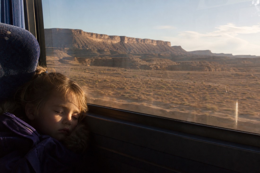 A young child asleep against a window seat on a tour coach crossing open desert in late afternoon light
