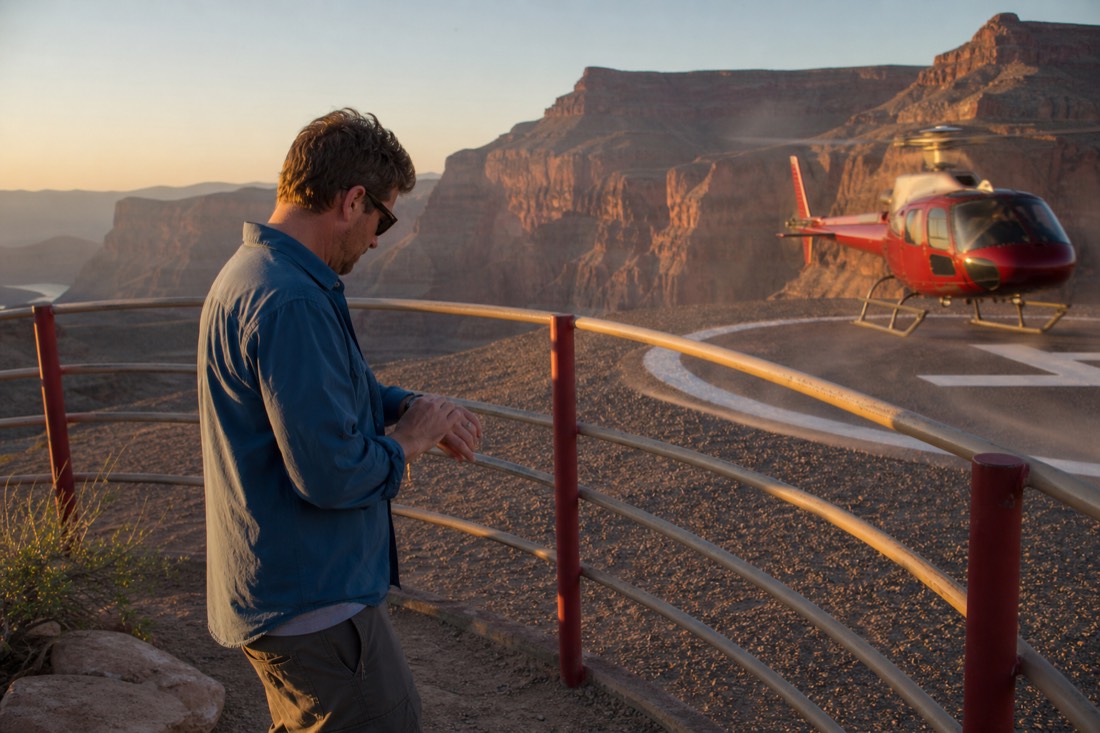 A traveler checking a watch beside a helipad fence as a tour helicopter spools up at golden hour