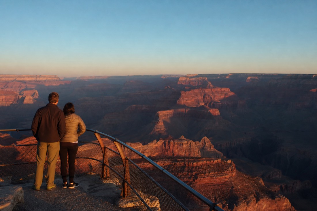 A couple standing at a South Rim overlook railing at first light with the canyon dropping away in layered shadow