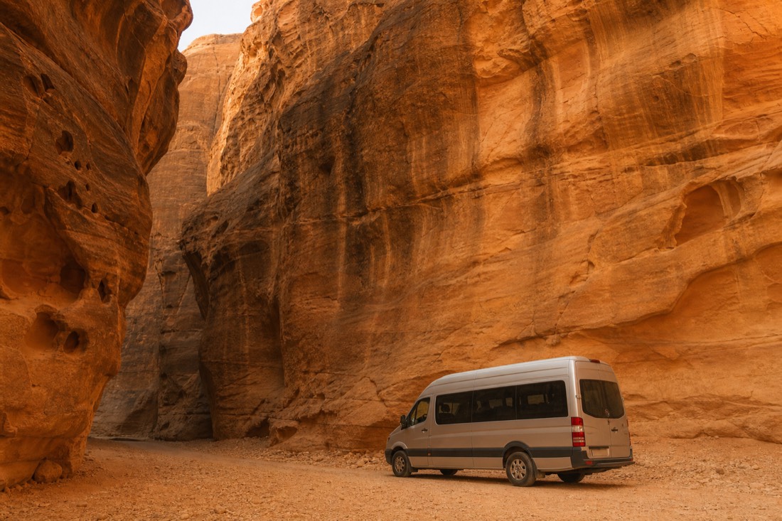 A small tour van parked beneath the sandstone walls of a slot canyon entrance with warm reflected light