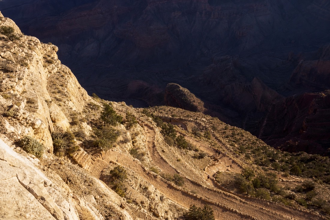 Steep switchbacks of a canyon trail descending into deep shadow below the rim with tiny hikers for scale