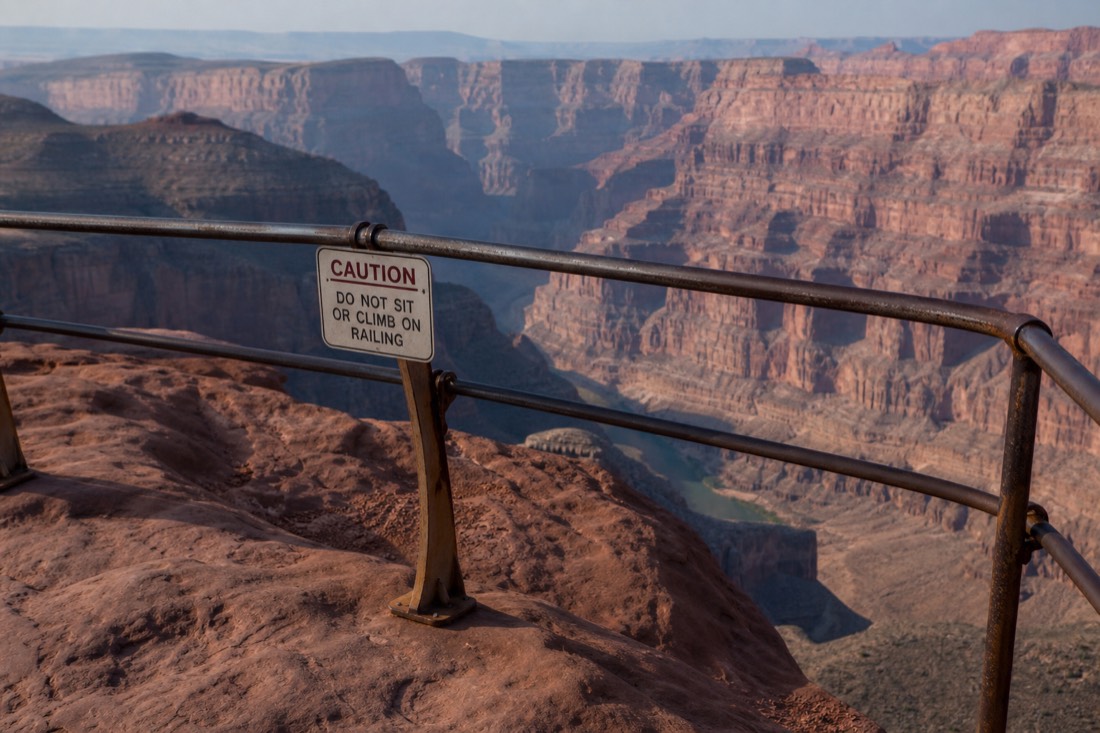 Steel railing along a rim overlook with warning signage and the sheer drop falling away beyond it