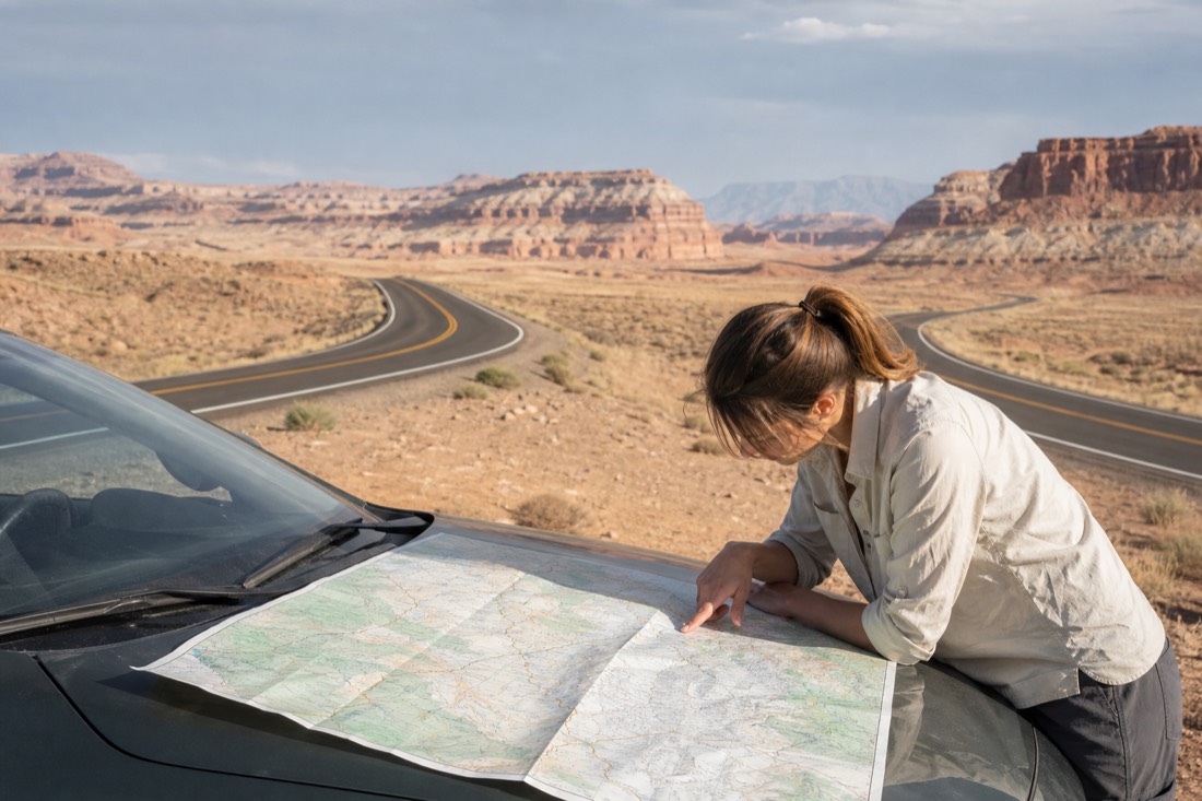 Traveler studying a paper road map on a car hood at a desert pullout with two highways splitting ahead